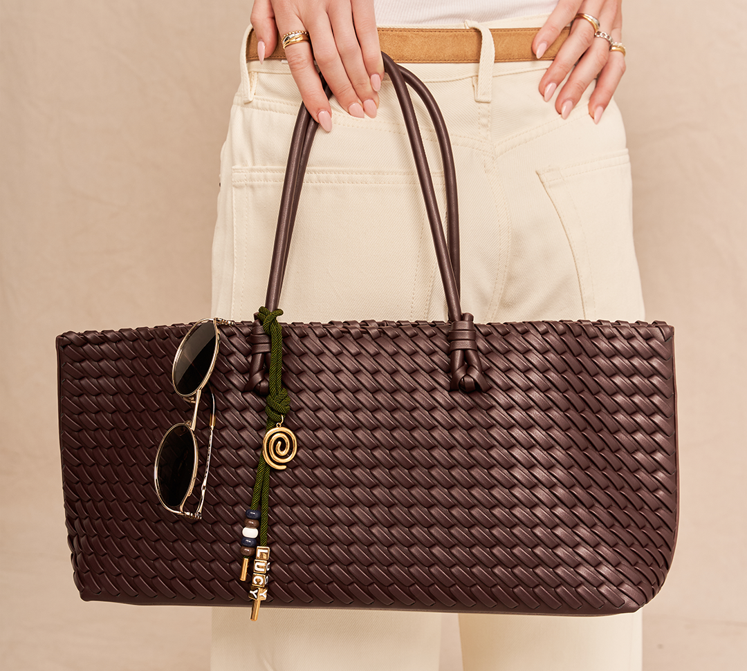 Brown woven handbag held by a person with sunglasses hanging from it against a beige background featuring a bag charm accessory