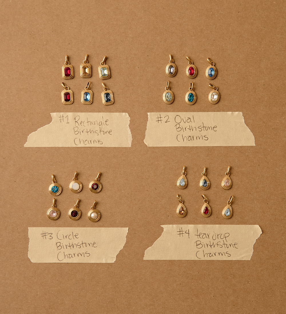 Four sets of small gold birthstone charms arranged on a brown kraft-paper background, with handwritten labels identifying the shapes: rectangular, oval, circle, and teardrop. Each set contains multiple charms in different birthstone colors.