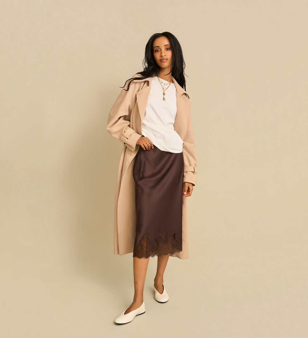 A woman models a necklace stack over a white top and a dark brown midi skirt paired with white flat shoes, against a solid beige background.
