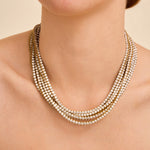 A close-up of a model wearing a multi-strand statement necklace with small round clear stones set in gold-toned metal, shown against a warm beige background.