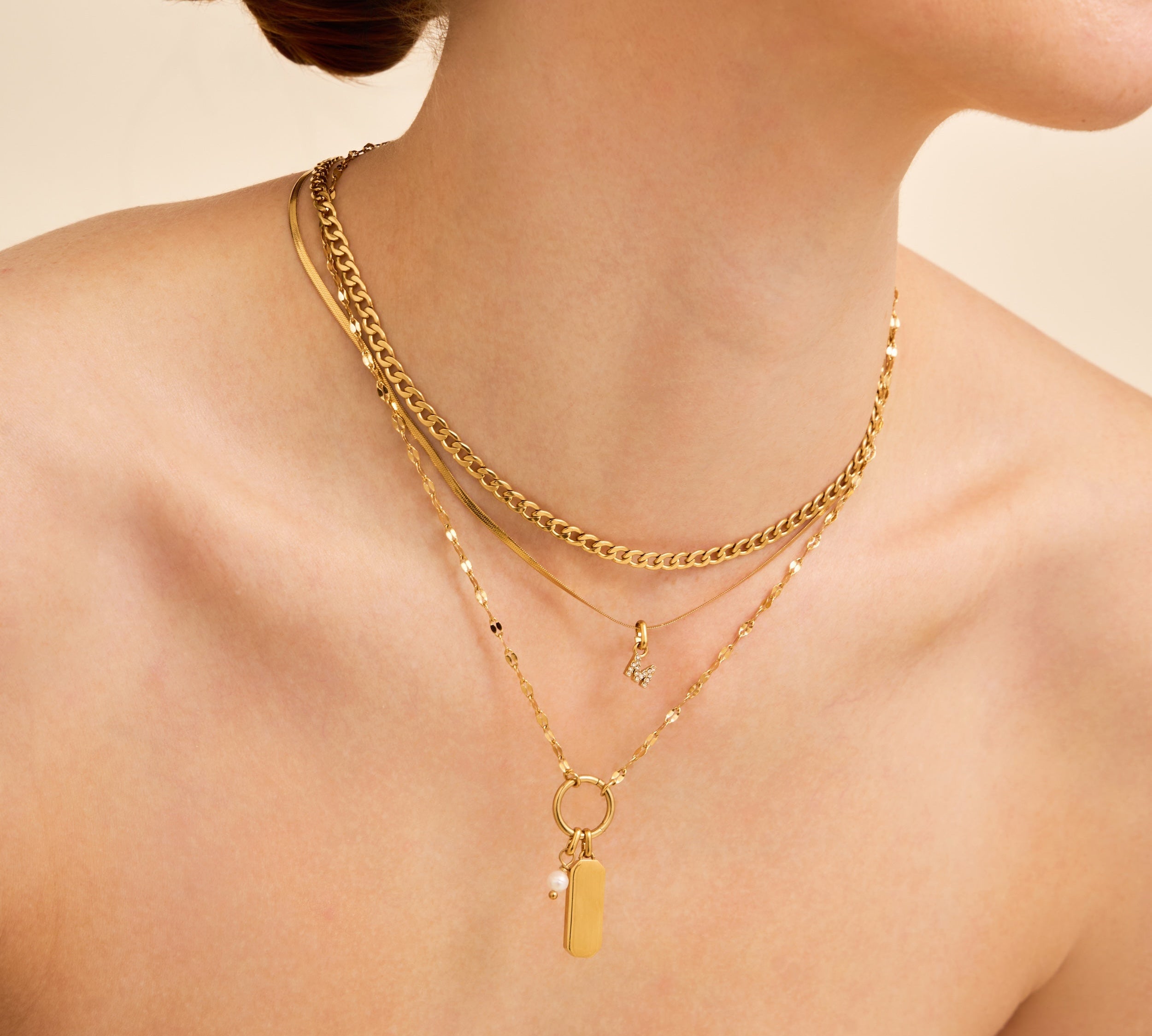 A close-up of a model wearing layered gold necklaces, including a curb chain, a delicate beaded chain, and a pendant necklace with a small pearl and rectangular charm, against a warm beige background.