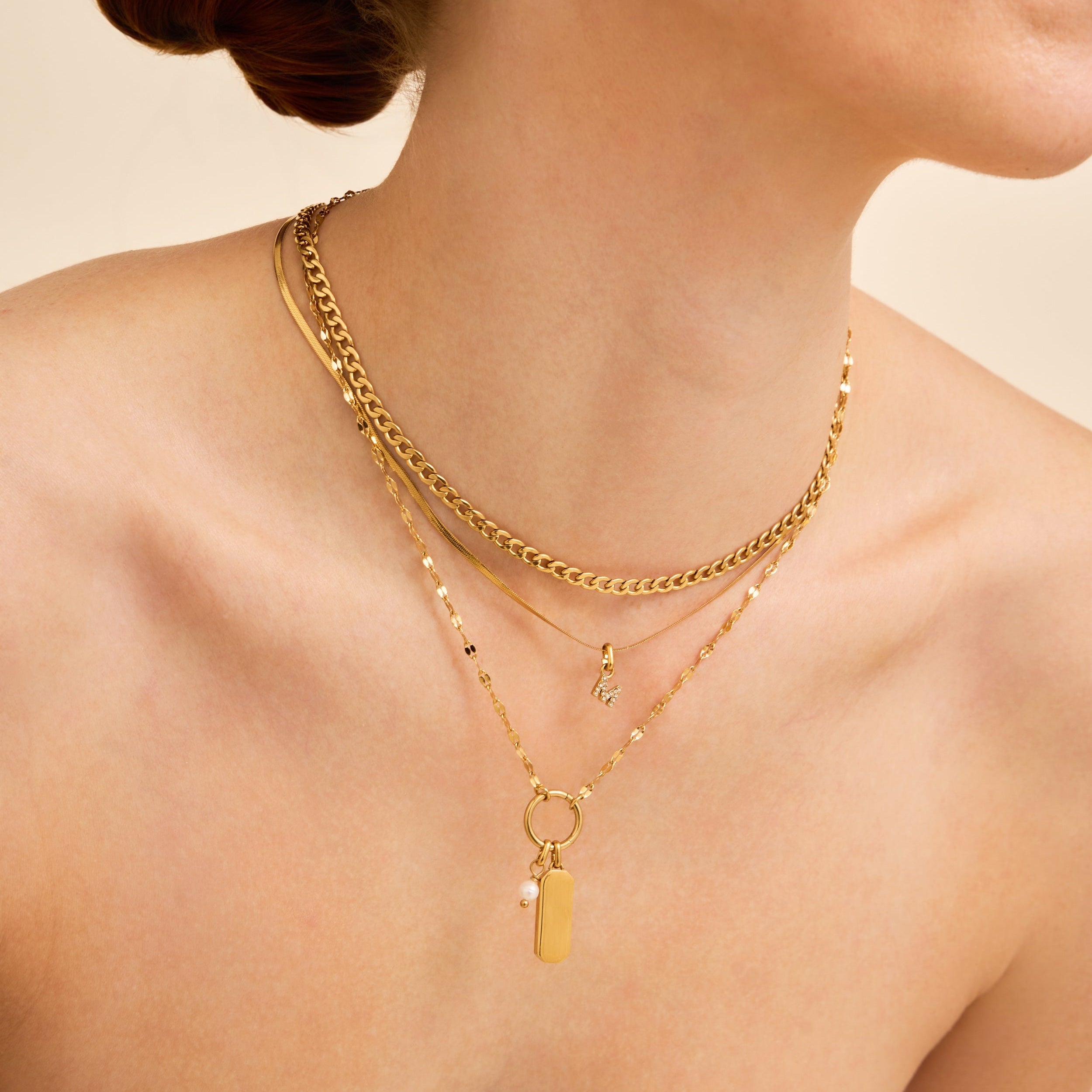 A close-up of a model wearing layered gold necklaces, including a curb chain, a delicate beaded chain, and a pendant necklace with a small pearl and rectangular charm, against a warm beige background.